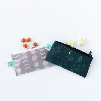 Two Bumkins Reusable Snack Bags, Small 2 Pack: Desert Night, are shown. One is gray with white moons, and the other is dark green with desert illustrations. Made from waterproof fabric, theyre surrounded by star-shaped orange crackers and cream-colored dinosaur snacks on a white background.