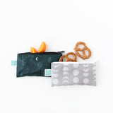 Two small Bumkins Reusable Snack Bags, Desert Night design, rest on a white surface. The top bag has a cactus and moon print and contains orange slices. The bottom bag is light gray with a geometric pattern and empty. Three pretzels are arranged to the right.