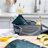 An open gray lunchbox sits on the kitchen counter with a banana visible inside, accompanied by sliced avocados and a Bumkins Reusable Snack Bag from the Small 2 Pack in Desert Night. The softly blurred background features a sink and kitchen items.