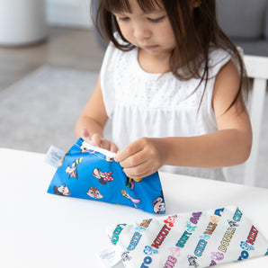 A young girl in a white dress opens Bumkins Reusable Snack Bag, Small 2 Pack: Mickey & Friends MVP at a white table.