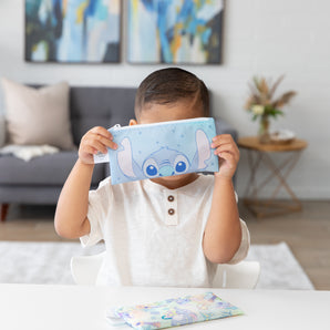 A child in a white shirt holds a Bumkins Stitch Star-ring Up Trouble snack bag at a table, with couch and abstract paintings behind.