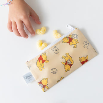 A child reaches for snacks near a Bumkins Reusable Snack Bag, Small 2 Pack: Pooh Bear and Friends on a white table.