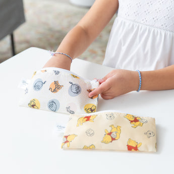 A child in a white dress places items into a Reusable Snack Bag, Small 2 Pack: Pooh Bear and Friends by Bumkins. Another bag with similar cartoon designs rests nearby. Made of waterproof fabric, these pouches are ideal for little adventures. The child dons blue bracelets on both wrists.