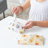 A child in a white sleeveless top puts a popsicle into a Bumkins Reusable Snack Bag, Small 2 Pack: Pooh Bear and Friends. A similar waterproof pouch lies nearby on the white table.