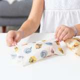 A child in a white dress gently holds a Bumkins Reusable Snack Bag, Small 2 Pack: Pooh Bear and Friends, featuring beloved Disney characters on waterproof fabric, resting on the childs hands against a white surface.