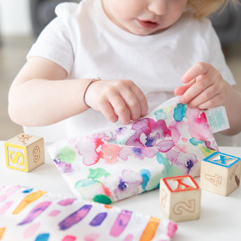 A child in a white shirt plays with colorful blocks and a Bumkins Reusable Snack Bag in Watercolor at the table.