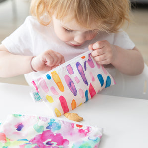 A child examines a Bumkins Reusable Snack Bag, Small 2-Pack: Watercolor & Brush Strokes, with a cookie in hand.