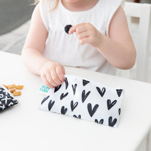 Child in white dress holding a snack above Bumkins Reusable Snack Bag (XOXO & Hearts) with cereal on the table.