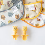 Two Winnie the Pooh baby bibs are laid out on a white surface, alongside yellow fork and spoon Silicone Chewtensils® by Bumkins, featuring playful character handles that add charm to baby-led weaning and make mealtime enjoyable.