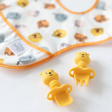 On a white surface, a colorful animal-patterned bib lays beside Bumkins Silicone Chewtensils®: Winnie The Pooh—a yellow bear-shaped spoon and fork set—ideal for baby-led weaning.