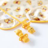 Two yellow Silicone Chewtensils® with a Winnie The Pooh bear theme rest on a white surface with sensory bumps for comfort. Behind them is a baby bib with a bear pattern and yellow trim, blending into the soft-focused background and enhancing the playful motif. Brand: Bumkins.