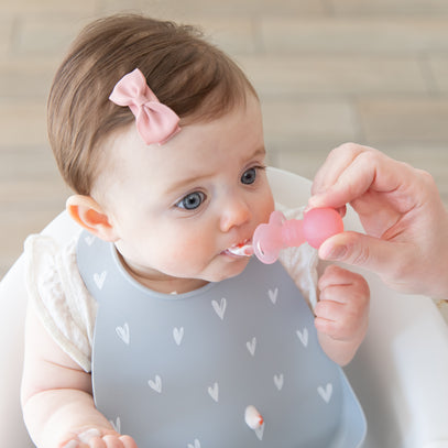 A baby with a pink bow in their hair sits in a high chair wearing a bib with heart patterns. They are being fed with a small pink spoon by an adult hand. The background is softly blurred.