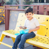 Young boy on a yellow bench, holding a Bumkins blue lunchbox with colorful toddler utensils, smiling brightly.