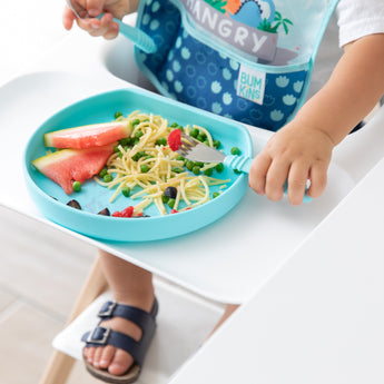 Child in a blue bib using Bumkins Spoon + Fork: Blue enjoys spaghetti, watermelon, and peas from a blue plate on their highchair.