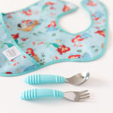 A blue bib with cartoon characters, paired with Bumkins Spoon + Fork: Blue and a turquoise silicone self-feeding utensil set.