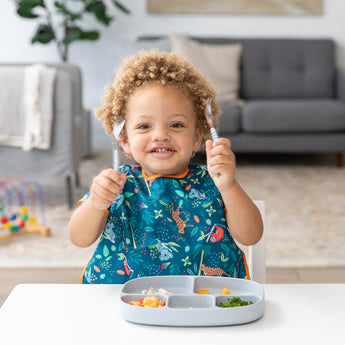 A curly-haired toddler smiles, holding Bumkins Spoon + Fork: Gray from their colorful utensil set at the table.