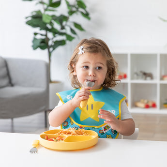 Toddler with a star bib uses Bumkins Pineapple Spoon + Fork, eating from a yellow plate; a potted plant and shelves in the background.