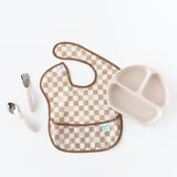 Checkered bib and cream plate paired with Bumkins Spoon + Fork: Sand, ideal for self-feeding on a pristine backdrop.