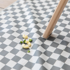 Tiny pasta and herbs sit on a Bumkins Splat Mat: Charcoal Check, next to a wooden table leg on the checkered floor.