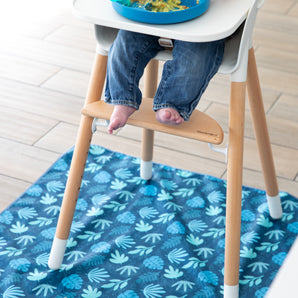 A baby in jeans sits barefoot on a wooden footrest of a high chair atop a Bumkins Blue Tropic splat mat with a leaf pattern.