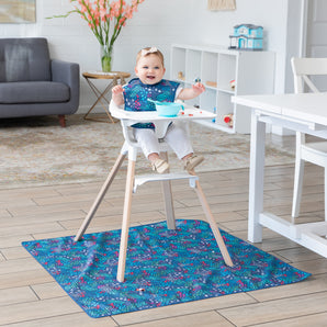 Baby in a highchair smiles with a blue bib, bowl on tray, and Bumkins Animal Prints Splat Mat underneath in the bright room.