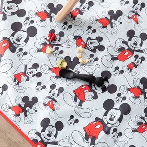 Bumkins Mickey Mouse Splat Mat features scattered food, a black spoon, and rolling pin on waterproof fabric. Machine washable for easy cleanup.