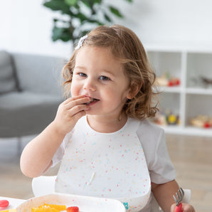 A toddler with curly hair eats at a table, wearing a Bumkins Silicone Bib: Vanilla Sprinkle and holding a fork, with fruit on the plate.