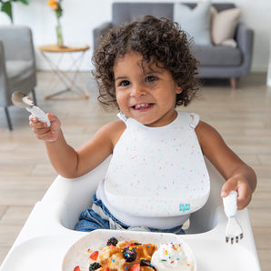 A curly-haired toddler beams in a high chair, gripping Bumkins Spoon + Fork: Vanilla Sprinkle, ready for fruit and waffles.