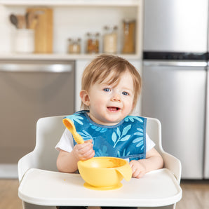 A baby in a Bumkins Starter Bib 2 Pack: Dinosaurs & Blue Tropic sits smiling with a spoon in a high chair against a kitchen backdrop.