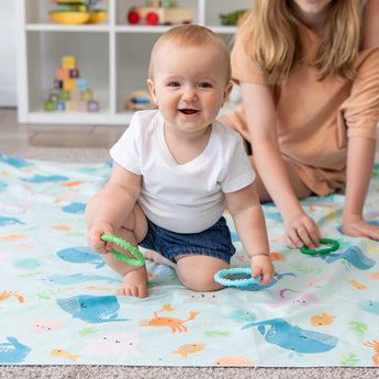 A baby on a sea creature play mat holds a Bumkins Silicone Teething Ring, while a woman in a peach shirt sits nearby.