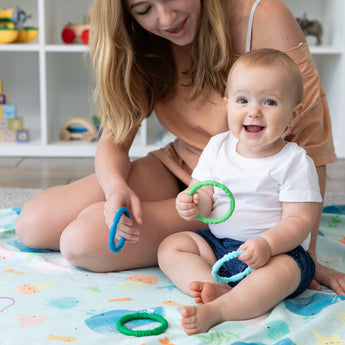 A baby plays with Bumkins Silicone Teething Rings on a playmat as a woman holds another, toys on shelves in the background.