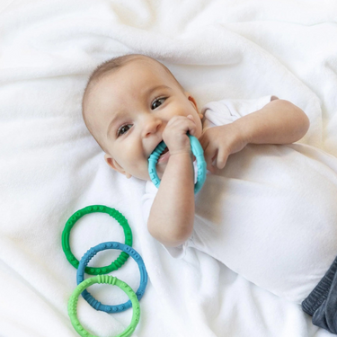 Baby on a white blanket chewing a Bumkins Summer Silicone Teething Ring, with green and blue rings nearby.