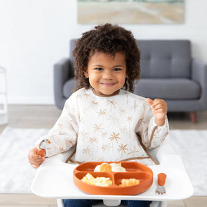 In a high chair, a curly-haired child smiles while holding a spoon. Theyre wearing the Bumkins Sleeved Bib: Palm Daze over a white shirt with palm tree patterns. An orange divided plate in front holds their baby-led weaning meal, and a gray couch is in the background.