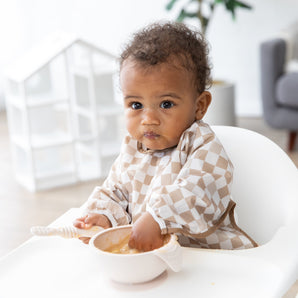 A baby in a high chair uses the Bumkins Sleeved Bib: Sand Check, trying baby-led weaning and wearing food cheerfully on their face.