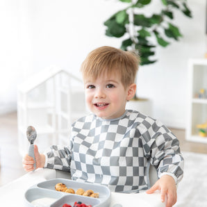 A child in a Bumkins Sleeved Bib: Charcoal Check sits at the table, smiling with a spoon, ready for baby-led weaning fun.