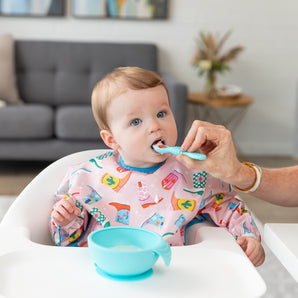 A baby in a high chair wears a Bumkins Sleeved Bib: Boots, being spoon-fed. A blue bowl sits on the tray with a sofa and plant behind.