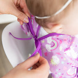 Adult hands tie the purple ribbon of a Bumkins Sleeved Bib: Unicorns, crafted from waterproof material with cloud and rainbow designs. A child in a white shirt sits in a white chair, ready for baby-led weaning.