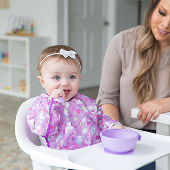 A baby wearing a Bumkins Sleeved Bib: Unicorns sits in a high chair, joyfully engaging in baby-led weaning with a spoon from a purple bowl. A smiling woman is nearby, and the well-lit room features shelves in the background.