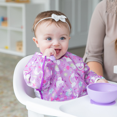 A baby in a high chair wears a Bumkins Sleeved Bib: Unicorns, enjoying self-feeding from a purple bowl while an adult is nearby.