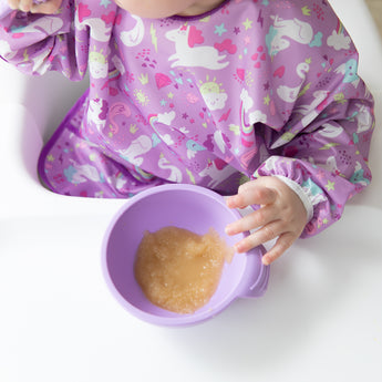 A baby in a high chair wears a Bumkins Sleeved Bib: Unicorns eagerly holds a lavender bowl of applesauce.