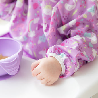 A child wears a Bumkins Sleeved Bib: Unicorns while sitting at a table, holding a lavender bowl filled with food.