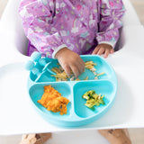 A toddler in a high chair wears a Bumkins Sleeved Bib: Unicorns made of waterproof fabric and holds a blue spoon. In front of them, a sectioned blue plate offers fries, mashed sweet potato, and sliced avocado, ideal for baby-led weaning adventures.