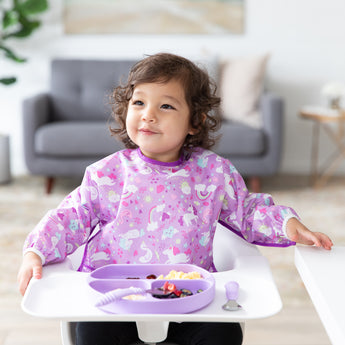 A curly-haired child sits in a high chair wearing a Bumkins Sleeved Bib: Unicorns made of waterproof fabric. A purple divided plate for baby-led weaning is on the tray. The background reveals a living room with a gray couch and lush plants.