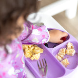 A child, wearing a vibrant Bumkins Sleeved Bib: Unicorns made from waterproof fabric, eagerly eats at the table. The purple plate, designed for baby-led weaning, is compartmentalized with cake crumbs and blueberries. A fork is neatly placed on the plate.