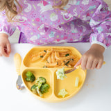 A child in a Bumkins Sleeved Bib: Unicorns enjoys a baby-led weaning meal with pasta, broccoli, and cucumber on a yellow divided plate, using a fork for cucumber pieces while a spoon lies beside the plate.