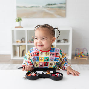 A toddler in Bumkins Sleeved Bib: Mickey & Friends Check Mates smiles at a table with food, surrounded by shelves of toys in the background.