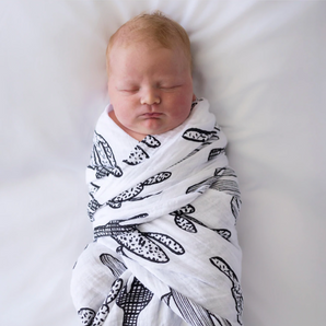 A newborn rests on a soft white surface in a high-contrast Modern Burlap Organic Swaddle, Cactus.