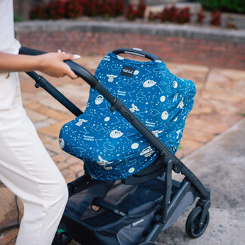 A person in a beige outfit strolls along a stone path, pushing a stroller covered with the Milk Snobs multifunctional Cover Star Wars The Child featuring space-themed patterns like rockets and planets, which also serves as a baby car seat and nursing cover.