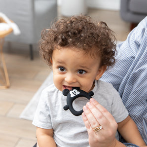 A curly-haired toddler bites a Bumkins Silicone Flat Teether: Mickey Mouse while sitting on an adults lap indoors.