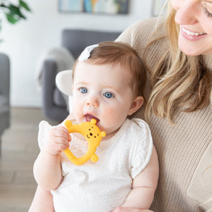 A baby with blue eyes chews on a Bumkins Silicone Flat Teether: Winnie the Pooh for teething relief, held by a smiling blonde woman. They are in a living room with a gray sofa and framed pictures in the background.